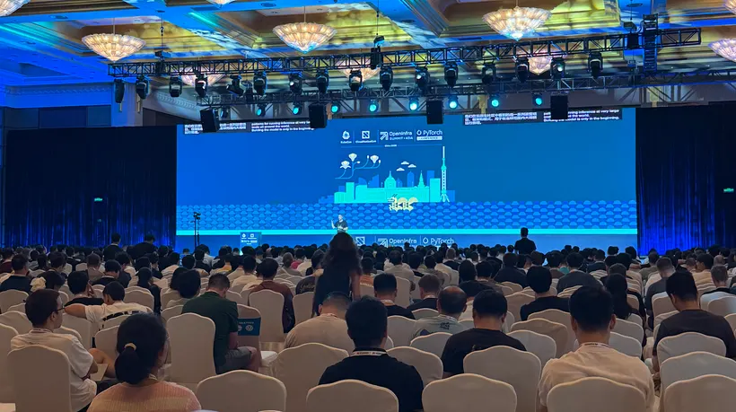 在上海举行的 KubeCon + CloudNativeCon + OpenInfra Summit + PyTorch Conference China 2026 开场主题演讲：舞台前坐满了人，其后的 LED 墙上是四个社区的 logo、"China 2026" 字样组合和插画风格的上海天际线，下方是一条双语实时字幕，写着 "patterns for running inference at very large scale all around the world"。