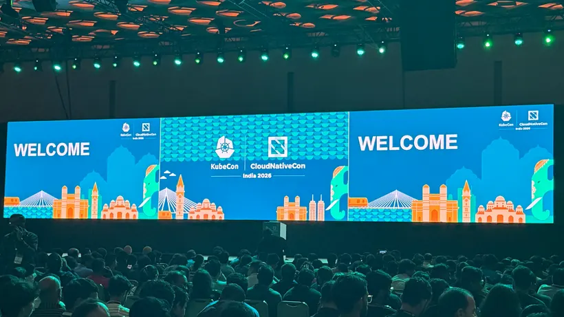 뭄바이에서 열린 KubeCon + CloudNativeCon India 2026의 가득 찬 오프닝 기조연설 현장: "WELCOME"과 행사 브랜딩, 그리고 일러스트로 그려진 뭄바이 스카이라인이 표시된 무대 LED 패널을 향해 객석을 가득 메운 청중